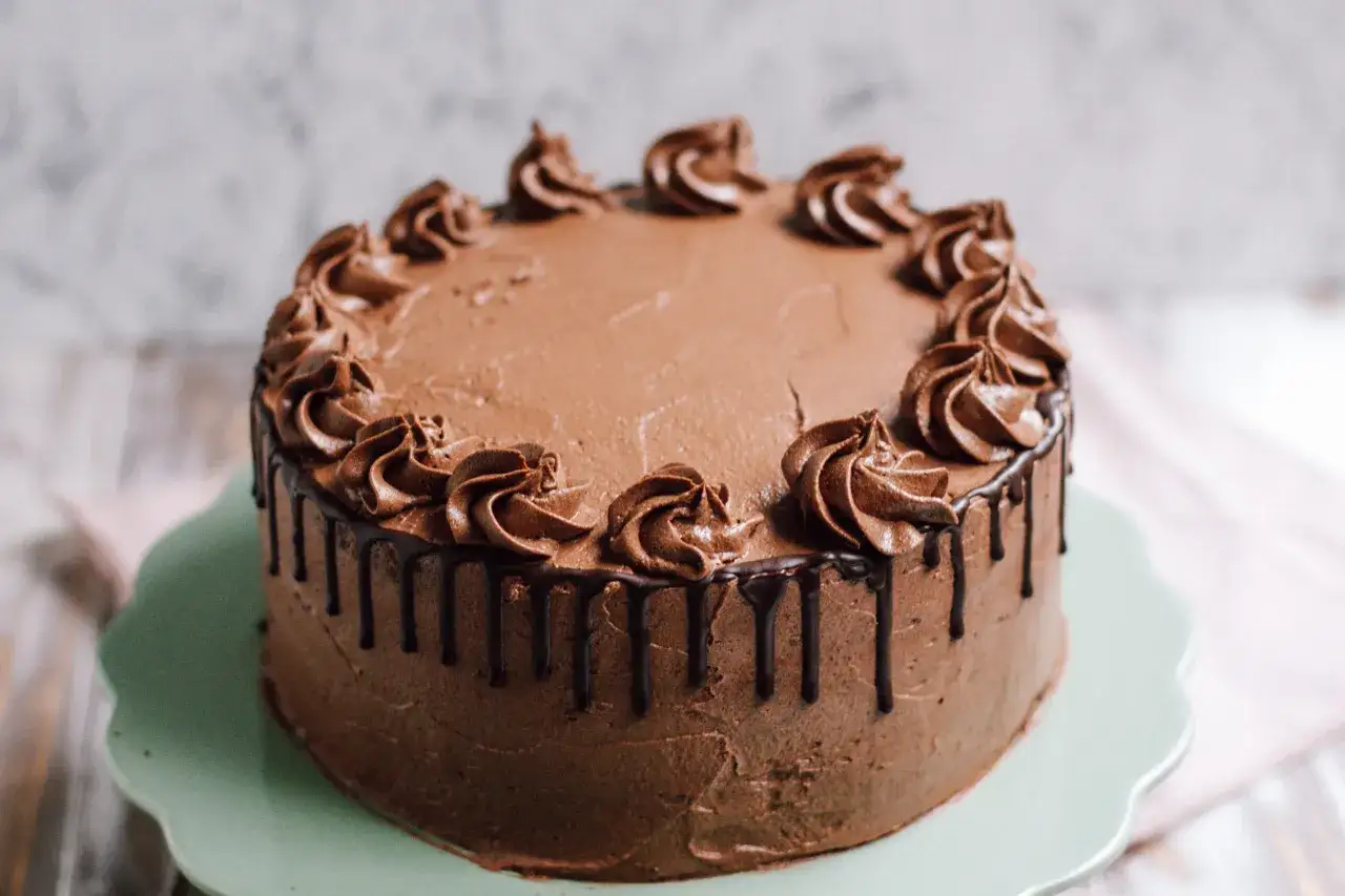 Eine k&ouml;stliche Schoko Torte mit Schokoladenglasur und Tupfen aus Schokoladencreme, die auf einem hellgr&uuml;nen Tortenst&auml;nder steht.