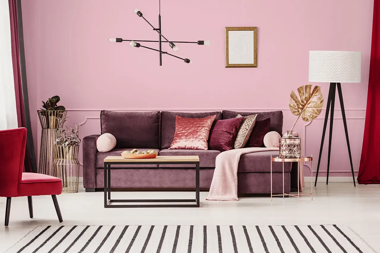 Elegancki salon w odcieniach brudny pudrowy róż i burgundu. Miękka sofa, czerwony fotel i złote dodatki tworzą przytulną atmosferę.