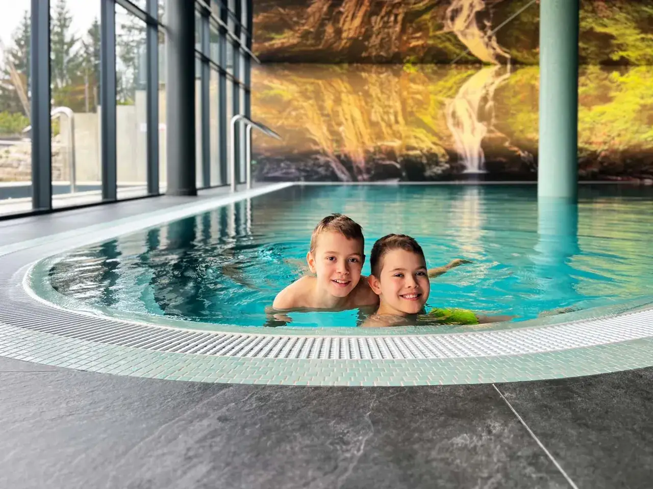 Dwóch uśmiechniętych chłopców pływa w basenie z widokiem na wodospad. Relaks w Złoty Horyzont Resort Szklarska Poręba.