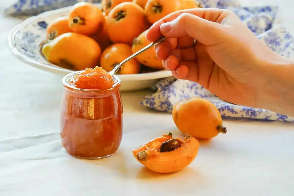 Un cucchiaino raccoglie la deliziosa marmellata di nespole da un vasetto, accanto a nespoli freschi e un piatto pieno.