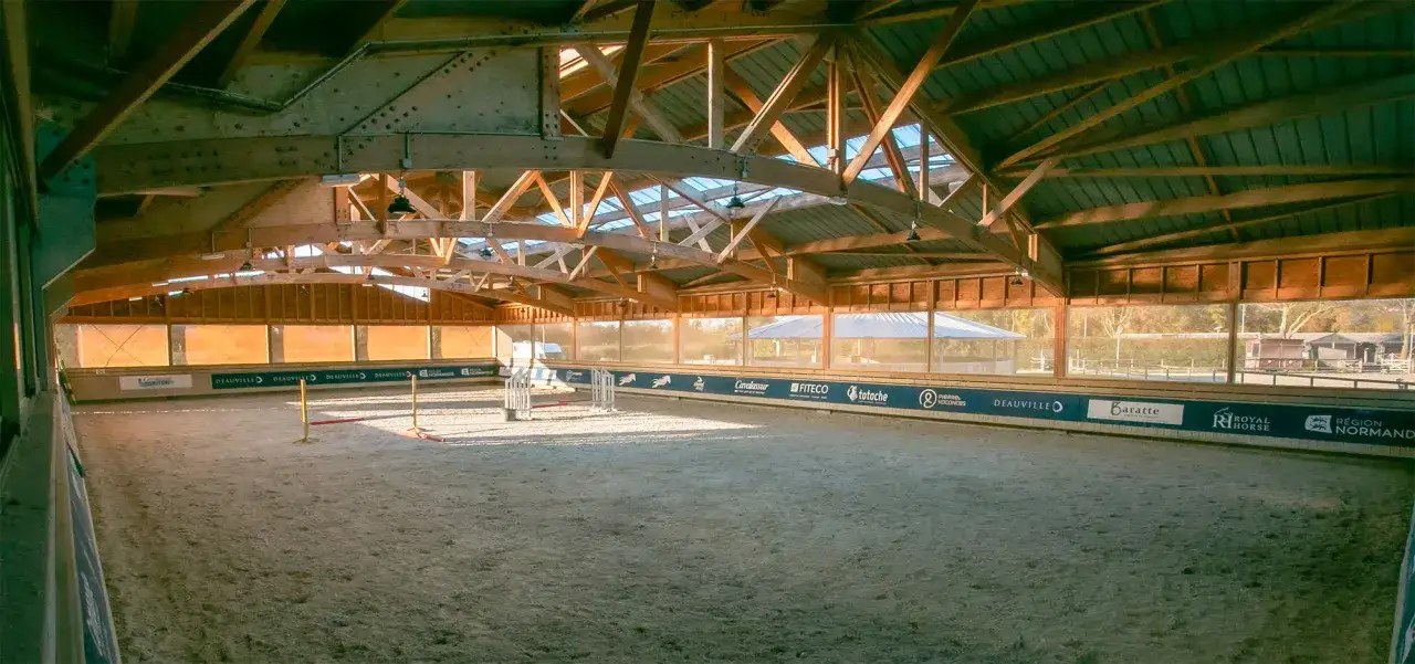 Intérieur d'une carrière de saut d'obstacle, avec une structure en bois et un toit vitré. Le sable est visible au sol, prêt pour l'entraînement.