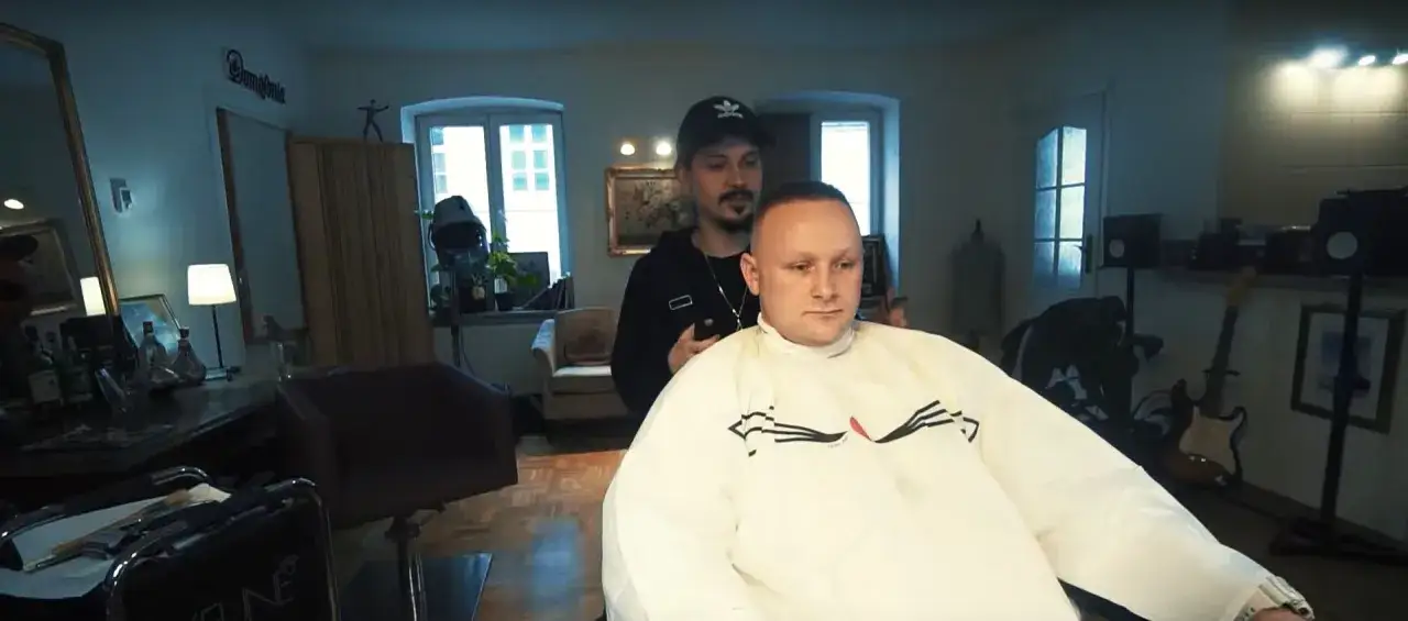 Fryzjer strzyże klienta w salonie. W tle widać gitary i lustro. Wiek Bajorsona nie jest widoczny, ale jego fryzura jest kr&oacute;tka.