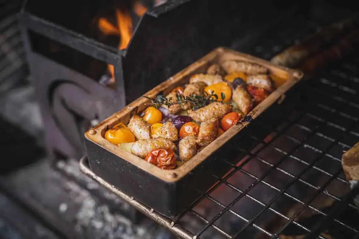 Salsiccia grillen in einer Keramikform mit Tomaten und Kr&auml;utern. Flammen lodern im Hintergrund.