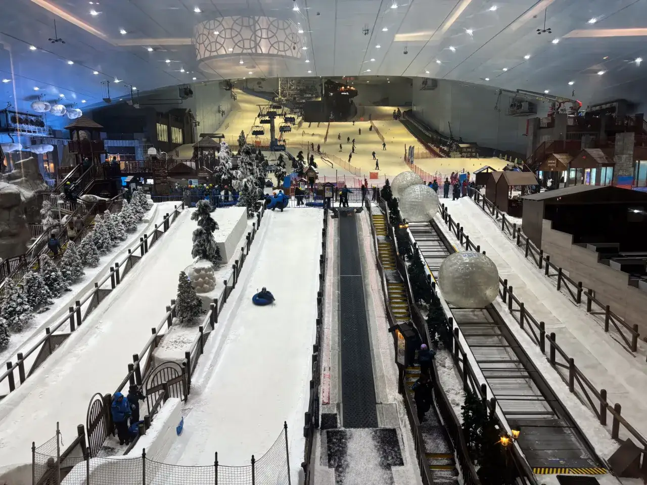 Wnętrze krytego stoku narciarskiego w Dubaju. Ludzie jeżdżą na nartach, snowboardach i zjeżdżają na pontonach. Widać wyciągi krzesełkowe i zjeżdżalnie.