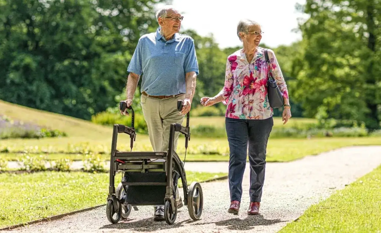 Ett äldre par promenerar på en grusväg i en park. Mannen använder en **rollator** för stöd.