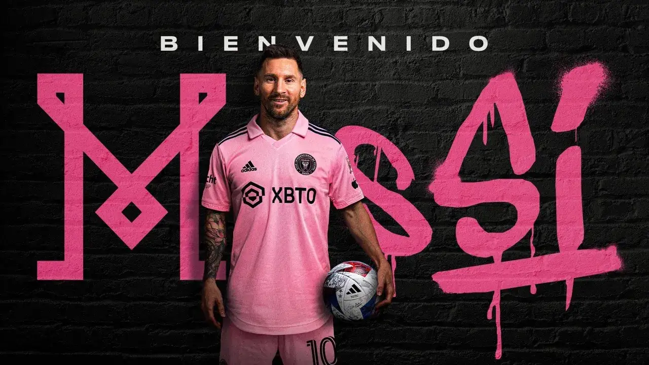 Lionel Messi w różowej koszulce Interu Miami, z piłką w ręku, na tle napisu
