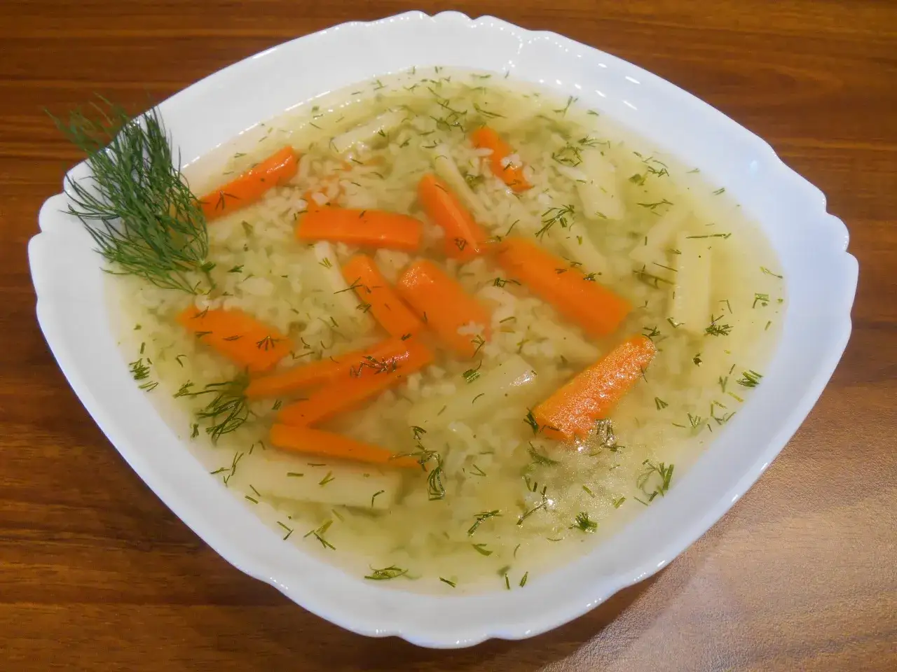 Aromatyczna zupa jarzynowa z marchewką, ryżem i koperkiem, podana w białej misce na drewnianym stole.