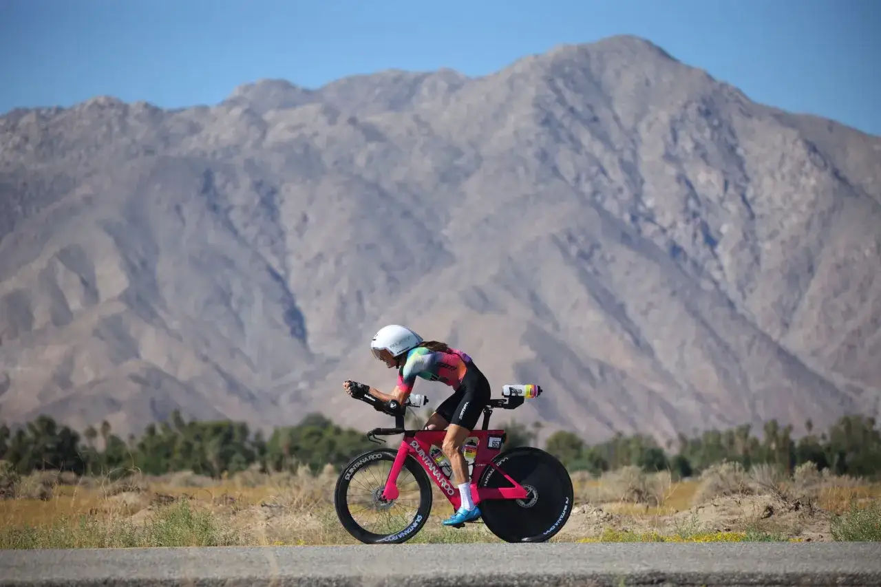 A cyclist races through a desert landscape, a scene straight out of a Tour de France fantasy. Mountains loom in the background under a clear blue sky.