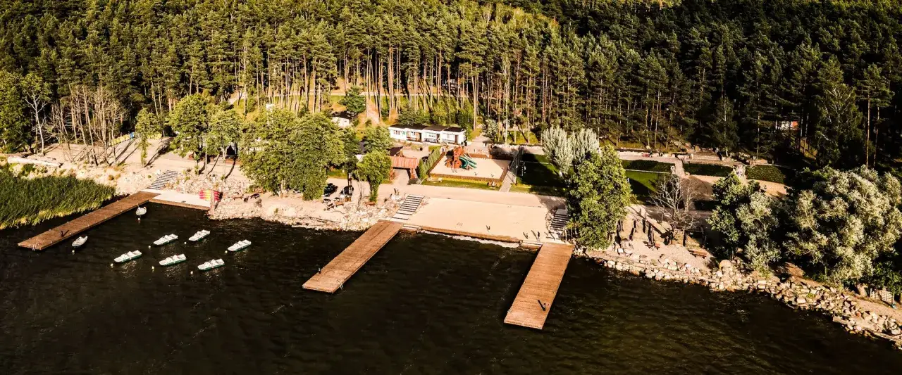 Ośrodek nad jeziorem z pomostami, plażą, placem zabaw i domkami wśr&oacute;d sosnowego lasu.