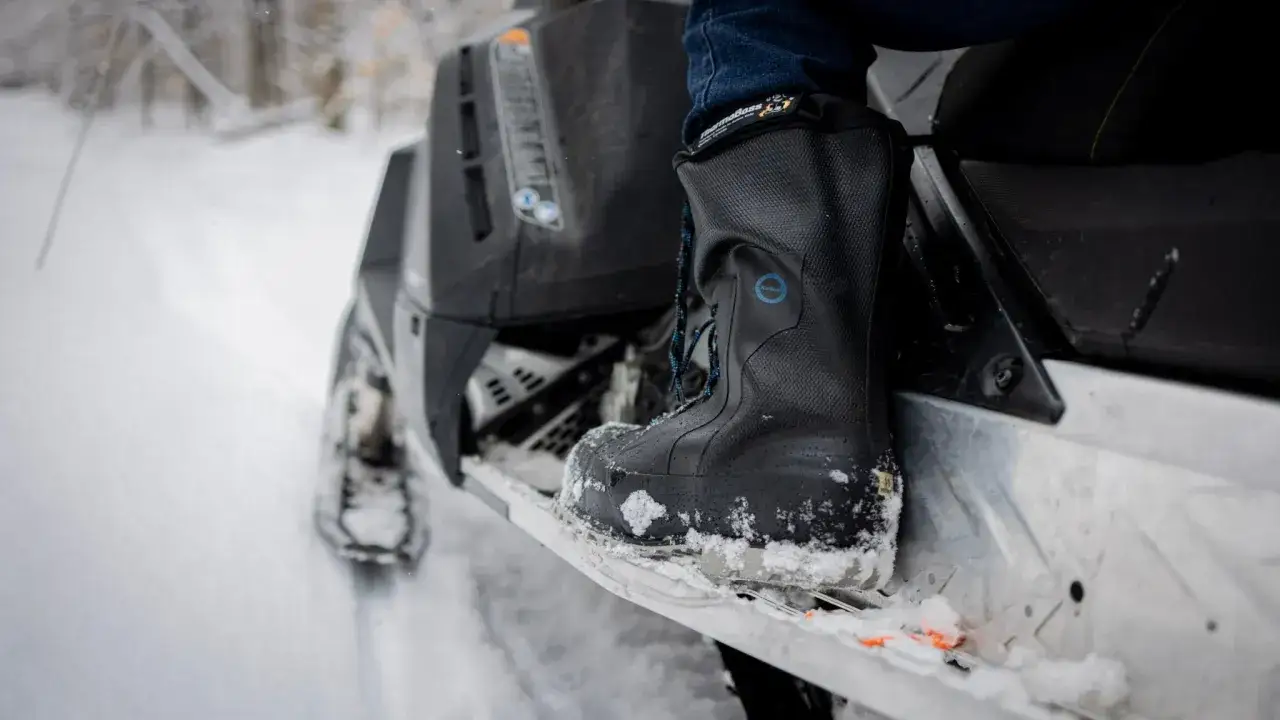 A person's black winter boot, showing how should winter boots fit snugly, rests on a snowmobile in a snowy landscape.