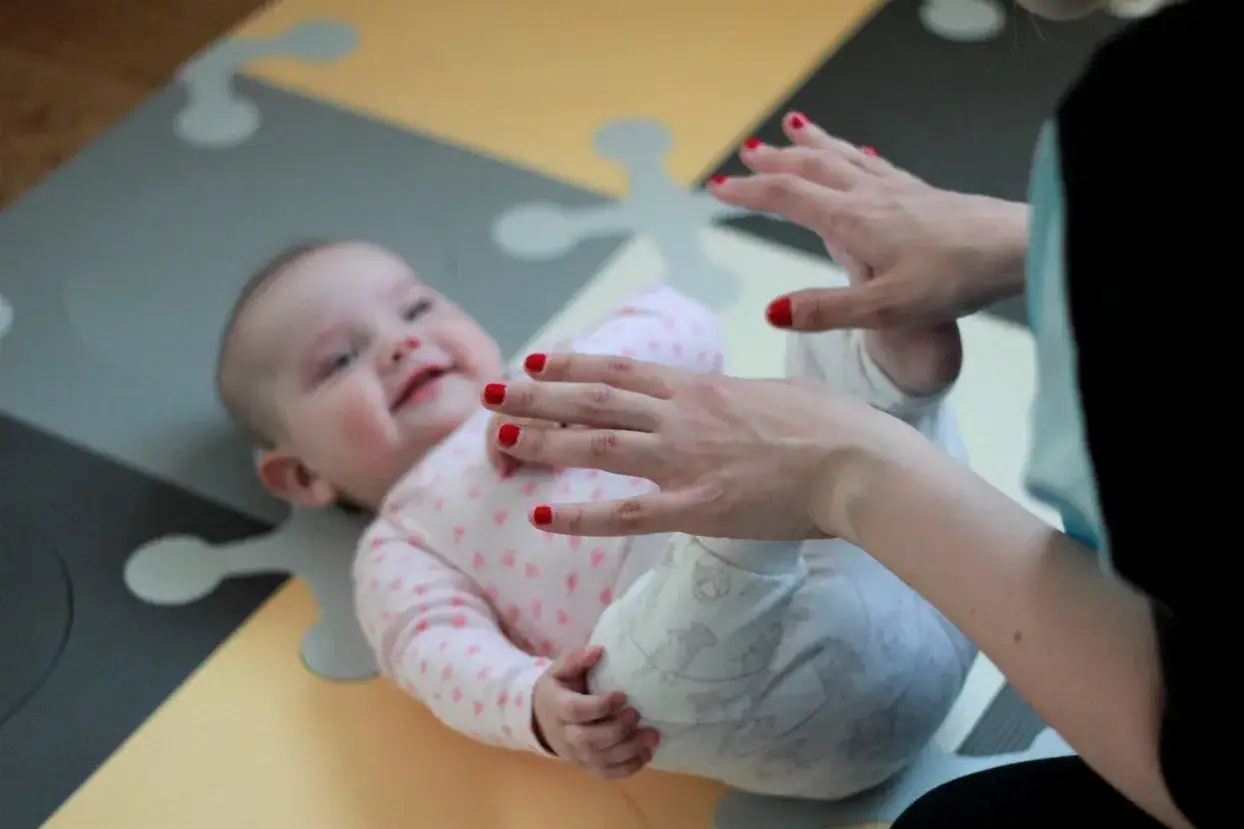 Uśmiechnięte 11 miesięczne dziecko leży na macie, a dorosła osoba z czerwonymi paznokciami delikatnie dotyka jego nóżek.
