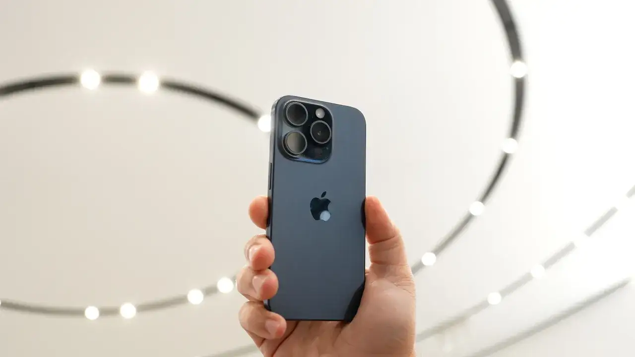 Main arri&egrave;re d'un iPhone 15 bleu nuit, tenu dans une main. Le logo Apple est visible, ainsi que le bloc de trois cam&eacute;ras.