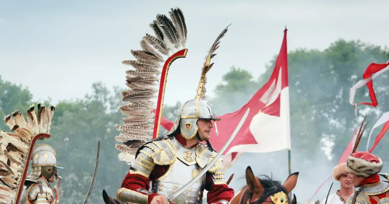 Huzar w zbroi i z pióropuszem na hełmie, z szablą w dłoni, na tle czerwono-białych flag.