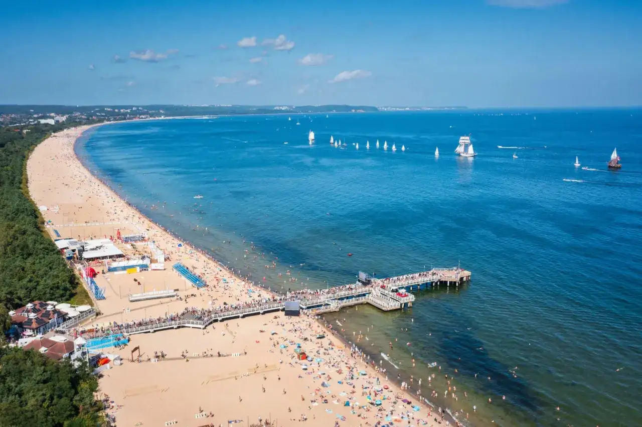 Plaża w Gdańsku pełna ludzi, molo, żagl&oacute;wki na morzu. Idealne miejsce, by sprawdzić, gdansk co robic latem.