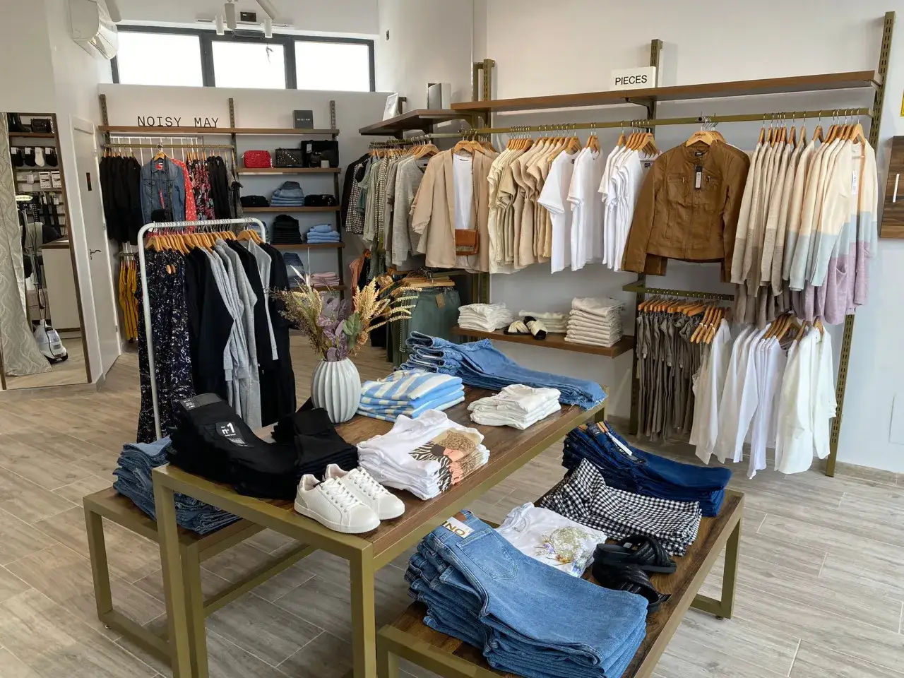 Interior de una tienda de ropa con percheros llenos de prendas y mesas con vaqueros y camisetas. ¡Todo listo para abrir una tienda!