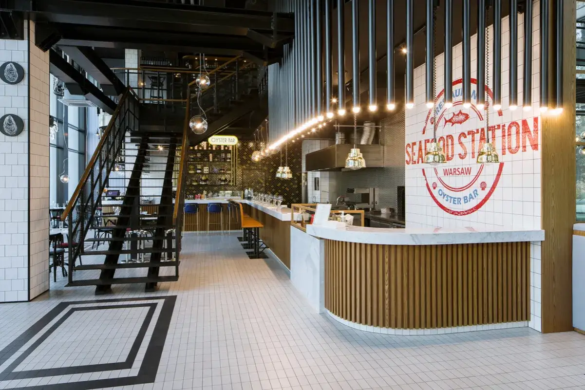 Nowoczesne wnętrze restauracji Seafood Station w Warszawie. Stylowy bar z białym blatem i drewnianą okładziną, a w tle schody i półki z butelkami.