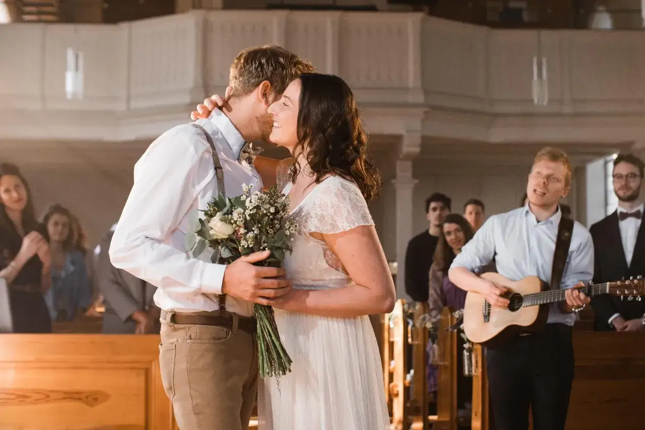 Hochzeitspaar umarmt sich, w&auml;hrend ein Mann Gitarre spielt und G&auml;ste zu bekannten Kirchenliedern zum Mitsingen lauschen.