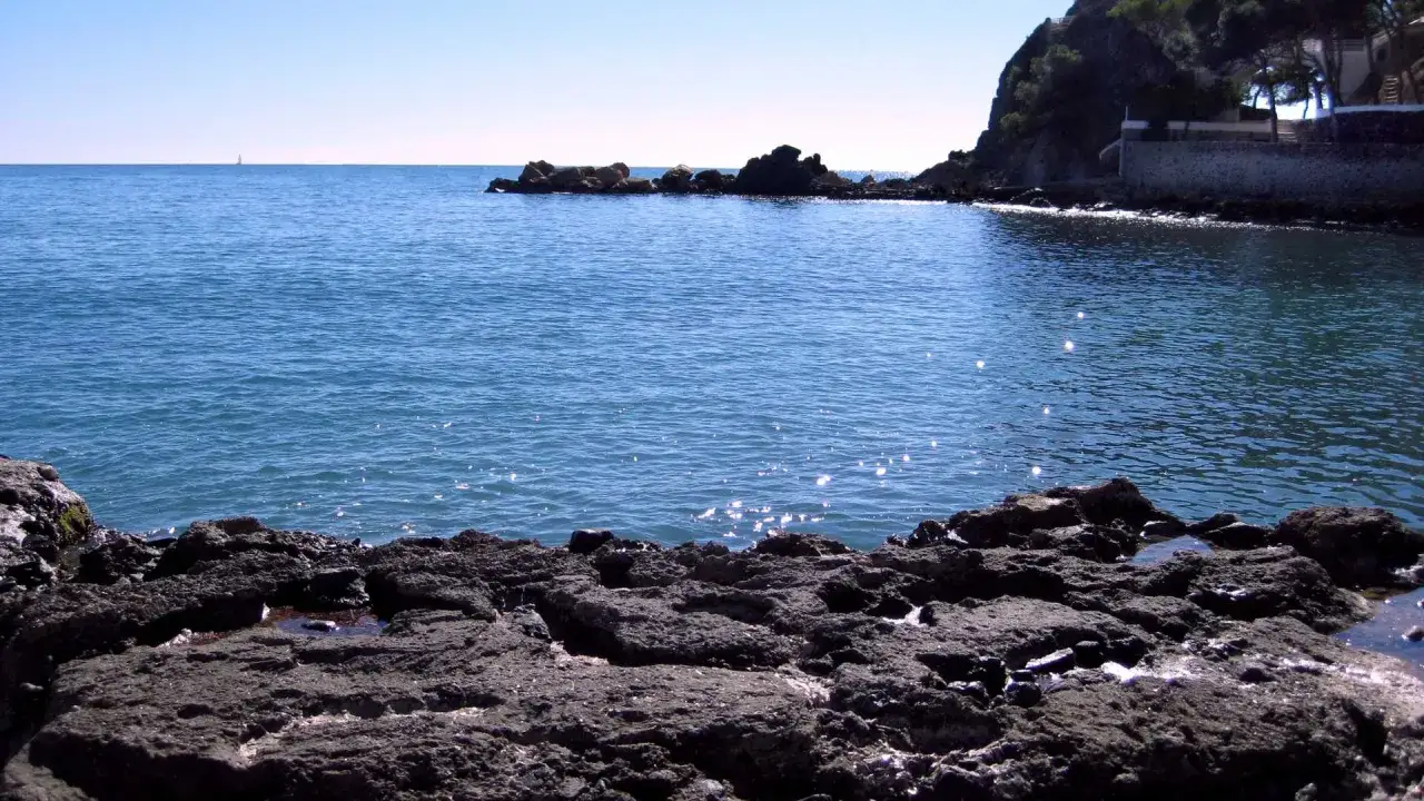 La cala del soio, con sus rocas oscuras y el mar azul brillante bajo un cielo despejado. Una vela solitaria en el horizonte.