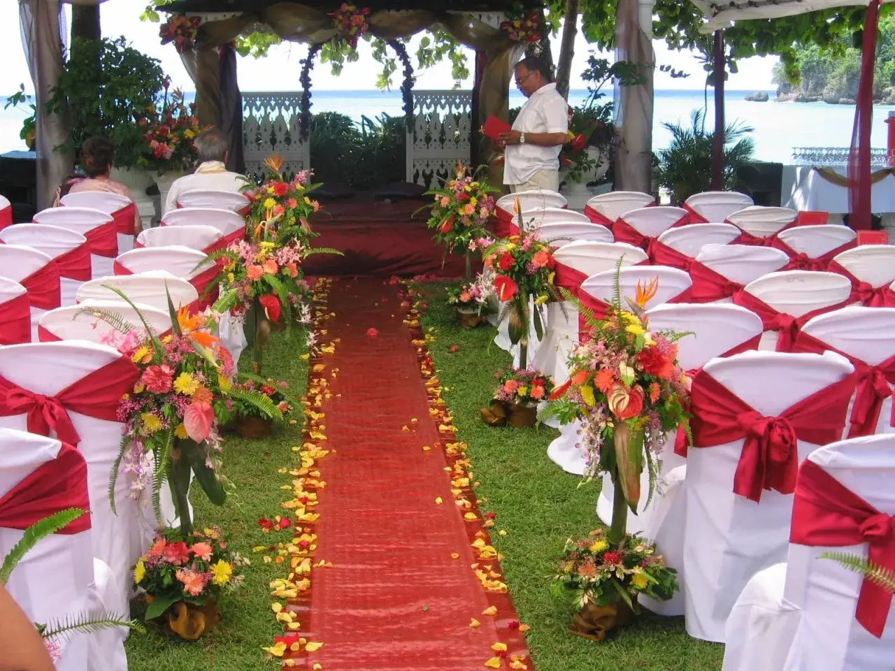 Allée de mariage thème Japon, décorée de fleurs tropicales et de pétales de roses, avec des chaises blanches ornées de rubans rouges.