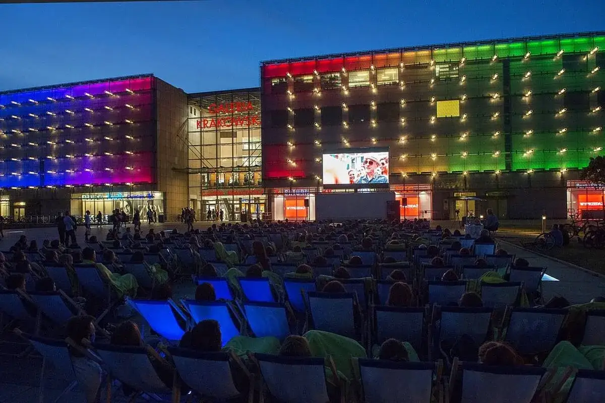 Wieczorne kino plenerowe przed Galerią Krakowską. Tłum widz&oacute;w na leżakach ogląda film wyświetlany na wielkim ekranie. Budynek galerii rozświetlony kolorowymi światłami.