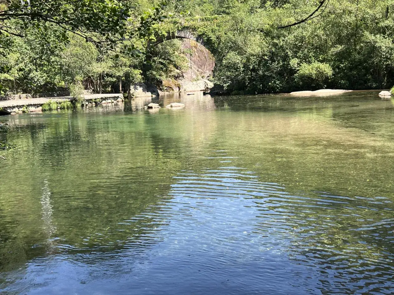 Aguas cristalinas en la playa fluvial Maceira, con un puente de piedra al fondo y vegetaci&oacute;n exuberante.