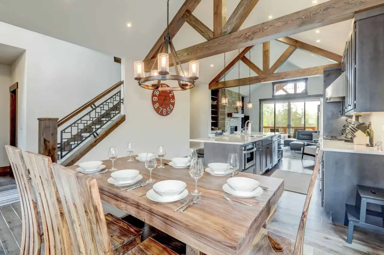 Stół z litego drewna i krzesła tworzą **modern rustic** klimat. W tle kuchnia z wyspą i salon z kominkiem.