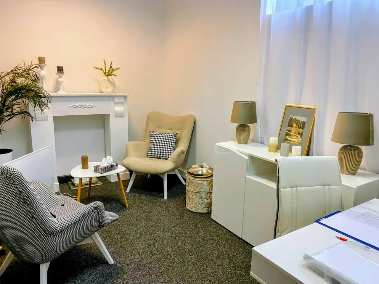 Przytulny gabinet terapeutyczny, idealny do sesji w trybie ambulatoryjnym. Dwa wygodne fotele, stolik z chusteczkami i świecą, kominek i lampy tworzą atmosferę spokoju.