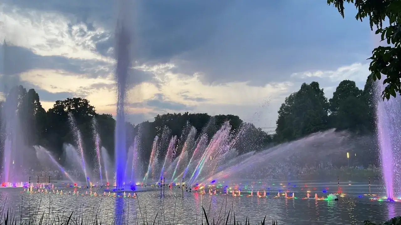 Spektakul&auml;re Wasserlichtkonzerte Fotos: Ein See mit bunten Font&auml;nen, die im Abendlicht tanzen, umgeben von B&auml;umen.