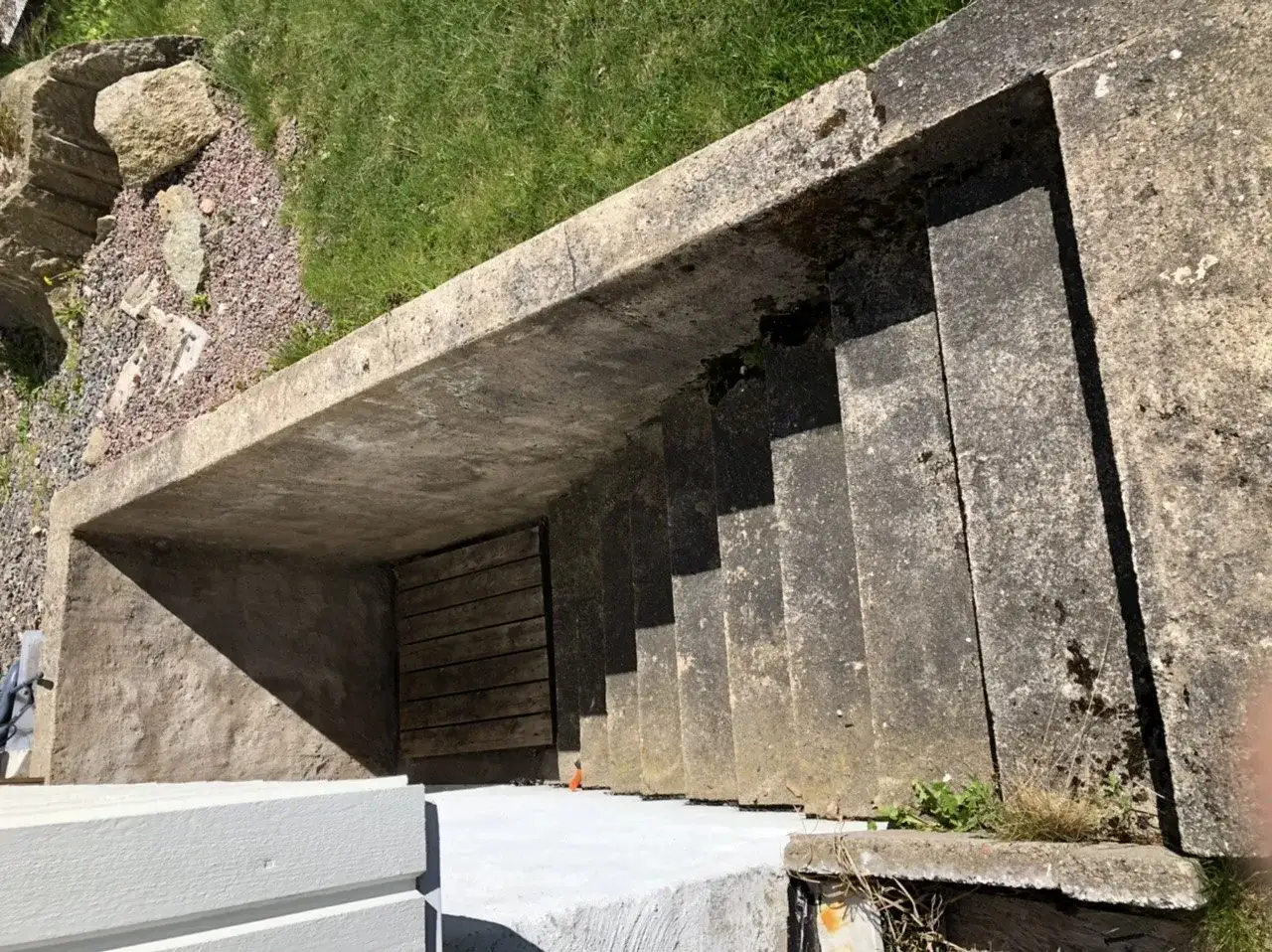 Betongtrappa leder ner till en träport. Gräs och stenar syns ovanför. Kanske någon ska borra i putsad fasad här?