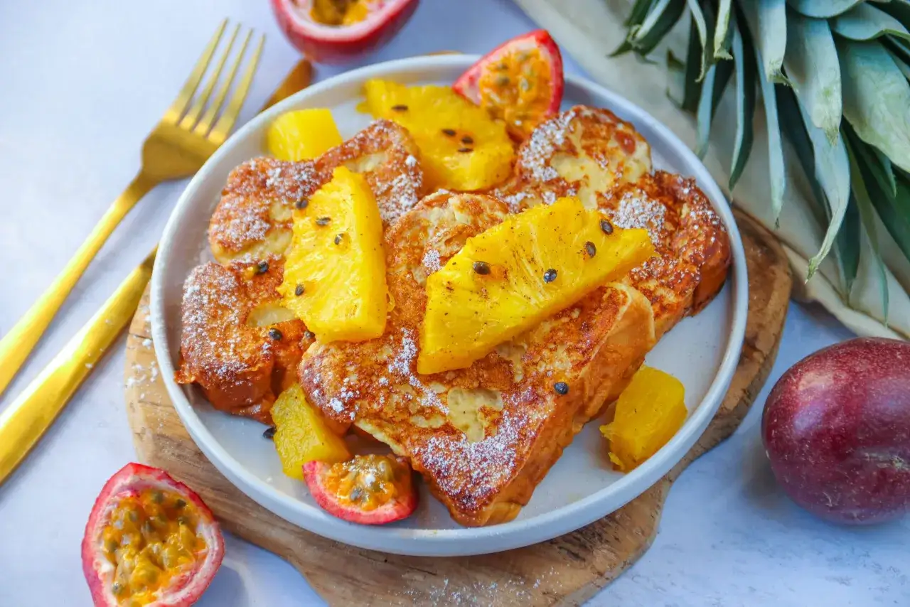 Une assiette de brioche perdue dorée, garnie de fruits de la passion et d'ananas frais, saupoudrée de sucre glace.