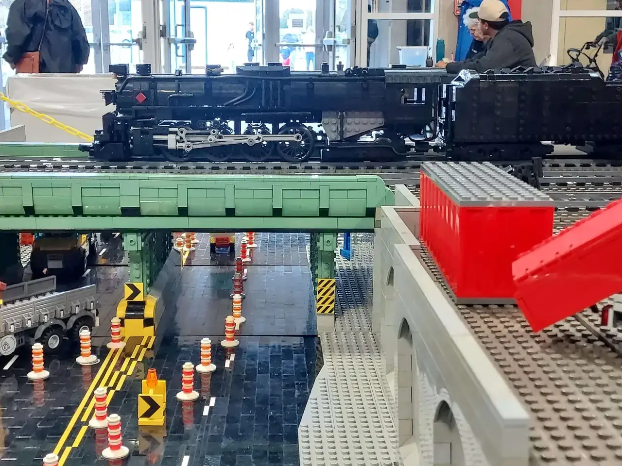 Beeindruckende Lego Dampflok MOC auf einer grünen Brücke über einer Straße mit Verkehrskegeln und einem LKW.