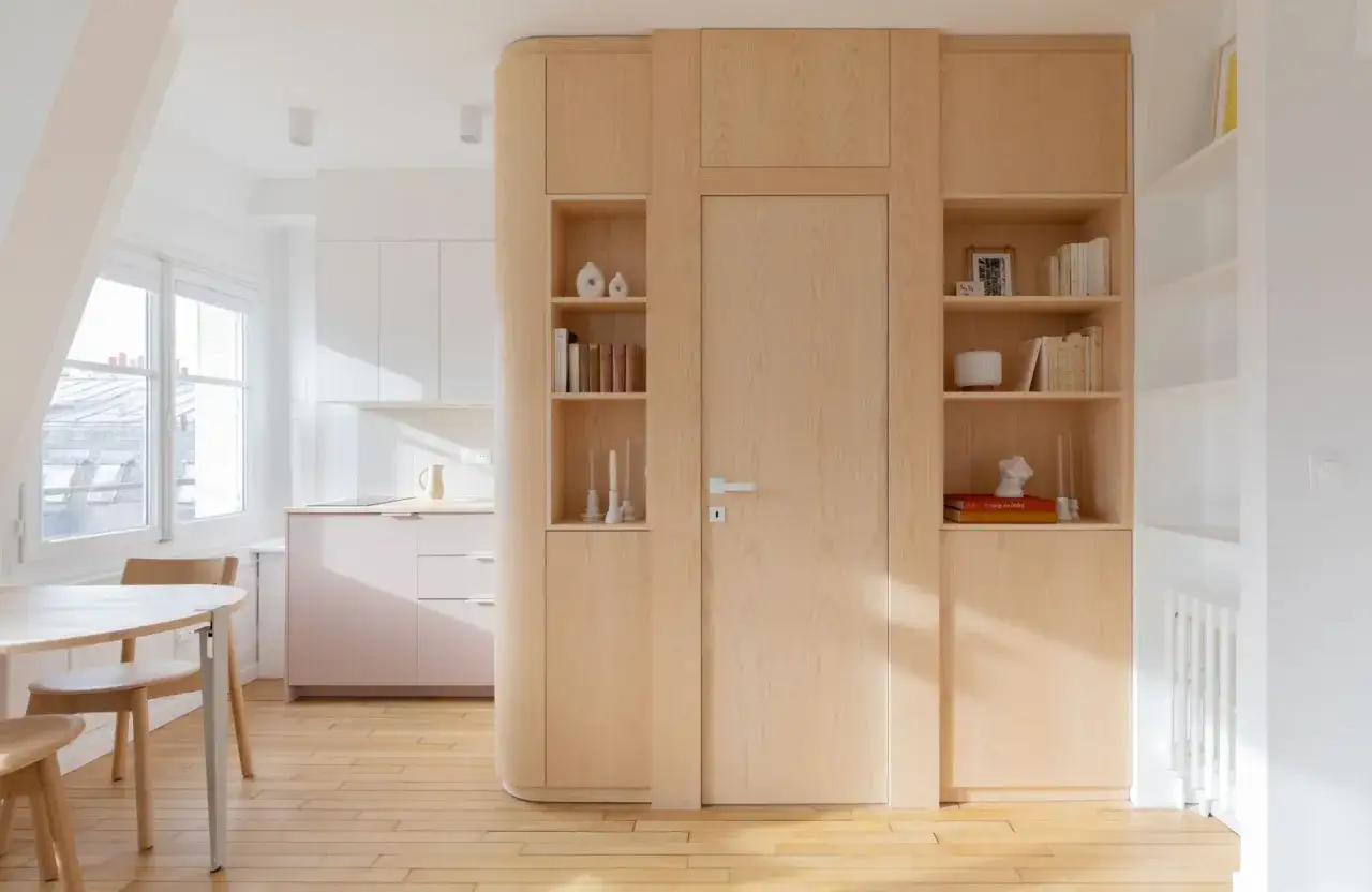 Am&eacute;nagement astucieux pour optimiser petit espace : cuisine rose p&acirc;le, salle &agrave; manger bois clair, et une grande biblioth&egrave;que int&eacute;gr&eacute;e autour d'une porte.