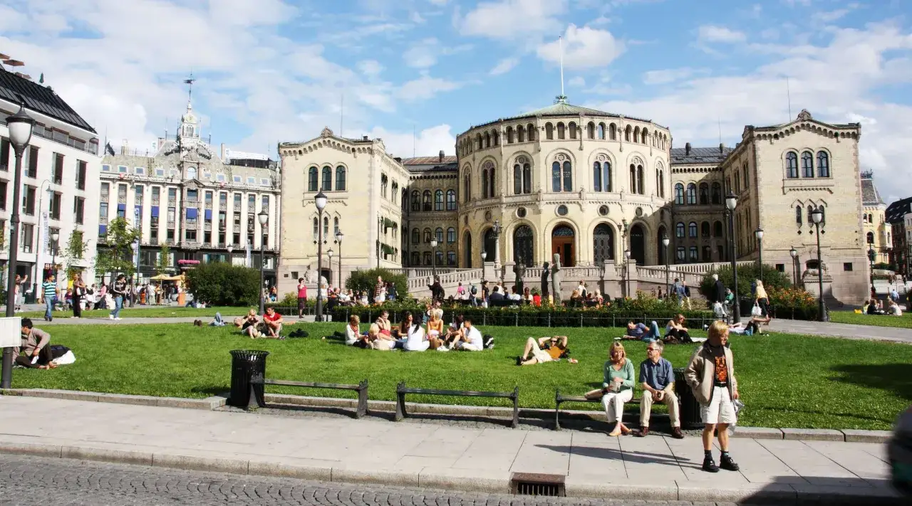 Ludzie odpoczywają na trawie przed budynkiem Stortinget, wzdłuż Karl Johans gate.