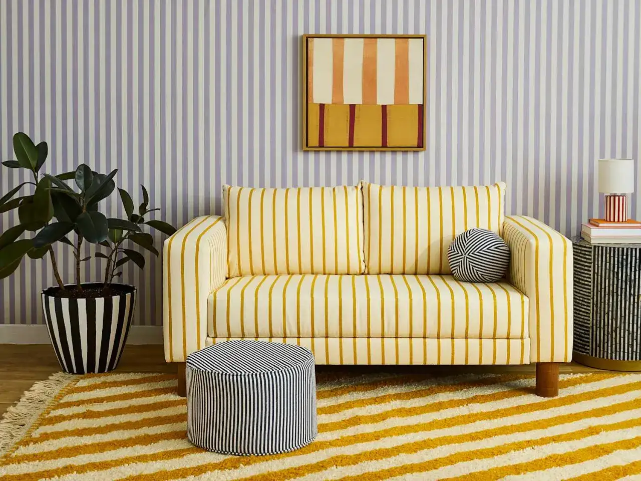 Un salon joyeux avec un canap&eacute; jaune &agrave; rayures, un pouf et un tapis assortis. Le papier peint violet &agrave; rayures compl&egrave;te le motif rayures.