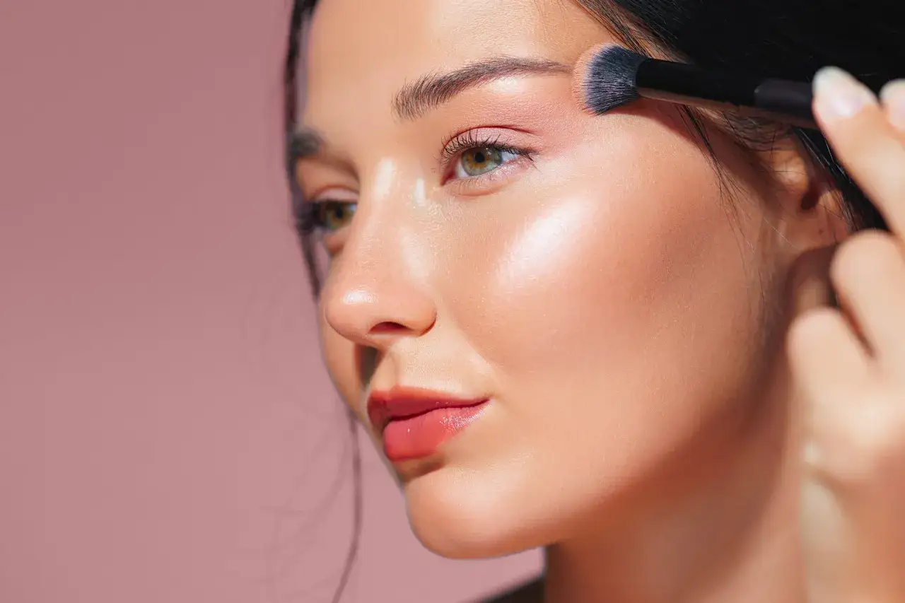 Une femme applique un fard à paupières rose pâle pour un maquillage yeux naturel. Sa peau est lumineuse et ses lèvres sont glossées.