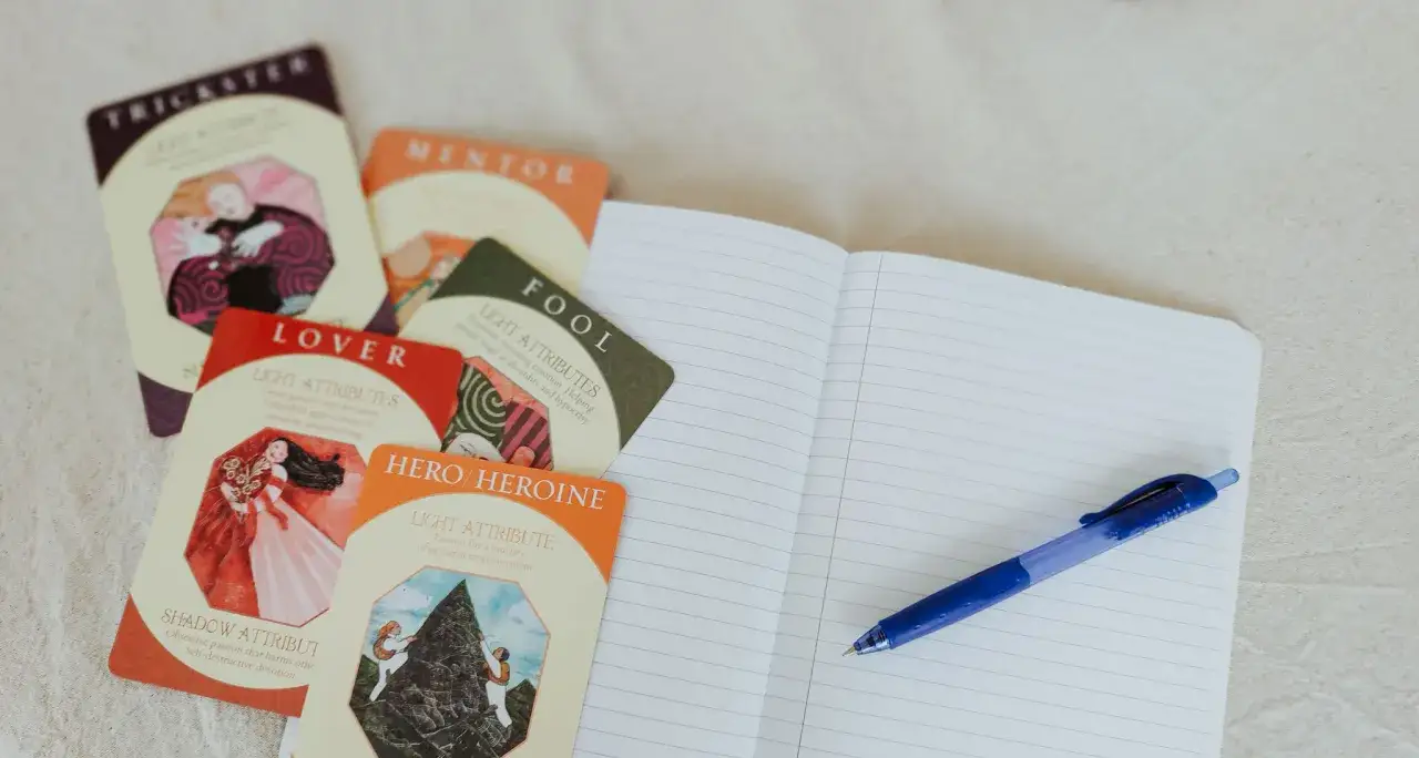 Cartes d'arch&eacute;types (Trickster, Mentor, Lover, Fool, Hero/Heroine) pr&egrave;s d'un carnet ouvert et d'un stylo bleu. Id&eacute;al pour explorer les personnages de roman.