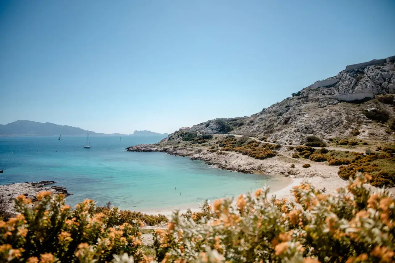 Vue panoramique d'une crique aux eaux turquoise sur la plage Frioul, avec des voiliers au loin et des collines rocheuses.