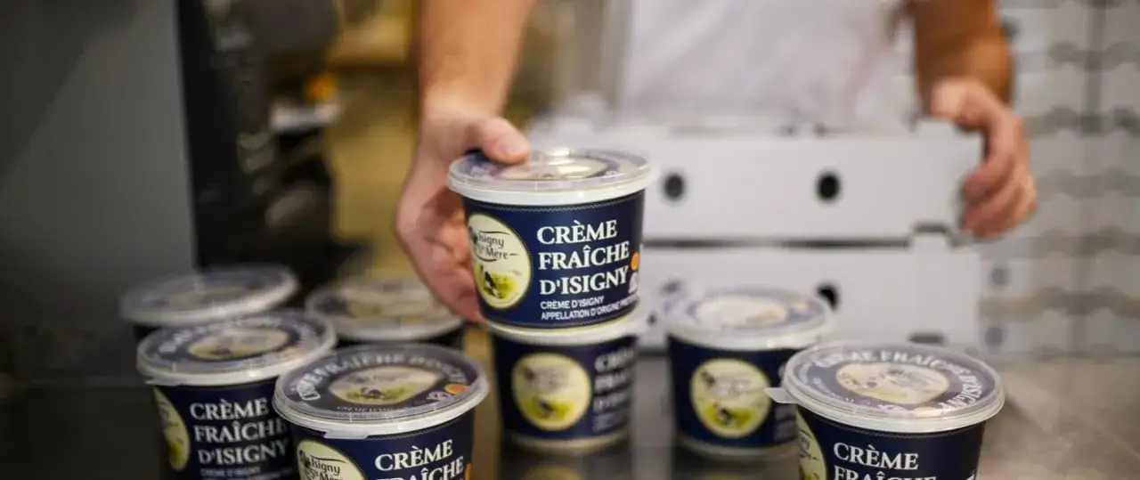 Main d'un ouvrier saisissant un pot de cr&egrave;me fra&icirc;che d'Isigny. Plusieurs pots sont align&eacute;s, pr&ecirc;ts &agrave; &ecirc;tre emball&eacute;s.
