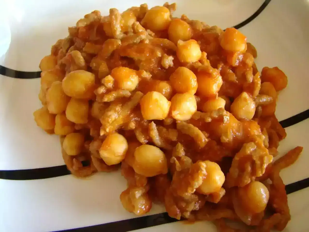 Un plato de garbanzos con carne picada, cocinado en una salsa rojiza y servido en una vajilla blanca con rayas negras.