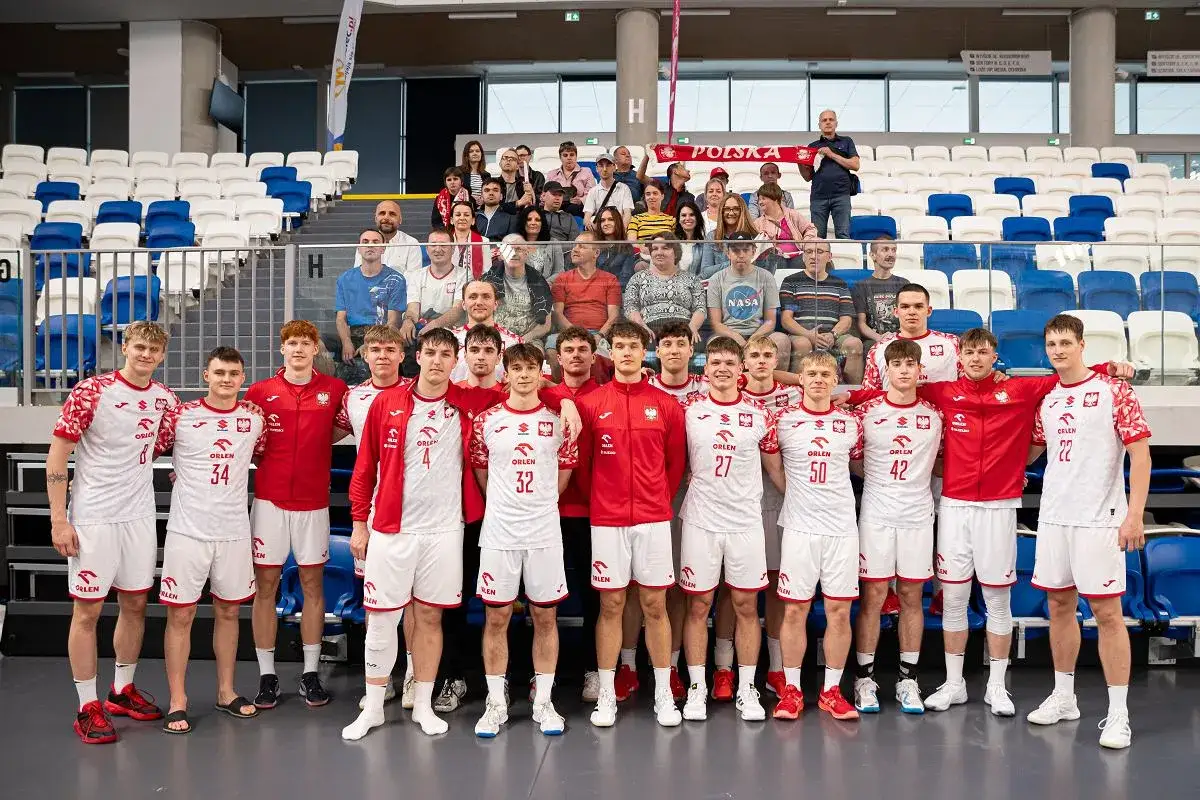 Młodzi piłkarze ręczni w strojach reprezentacji Polski pozują do zdjęcia z kibicami na trybunach.