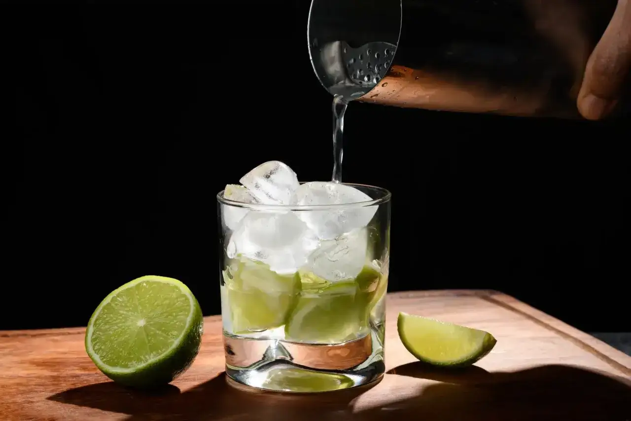 Eiswürfel und Limettenspalten in einem Glas, während eine Flüssigkeit aus einem Shaker gegossen wird. Das ist die Zubereitung eines Caipirinha, der mit Cachaca gemacht wird.