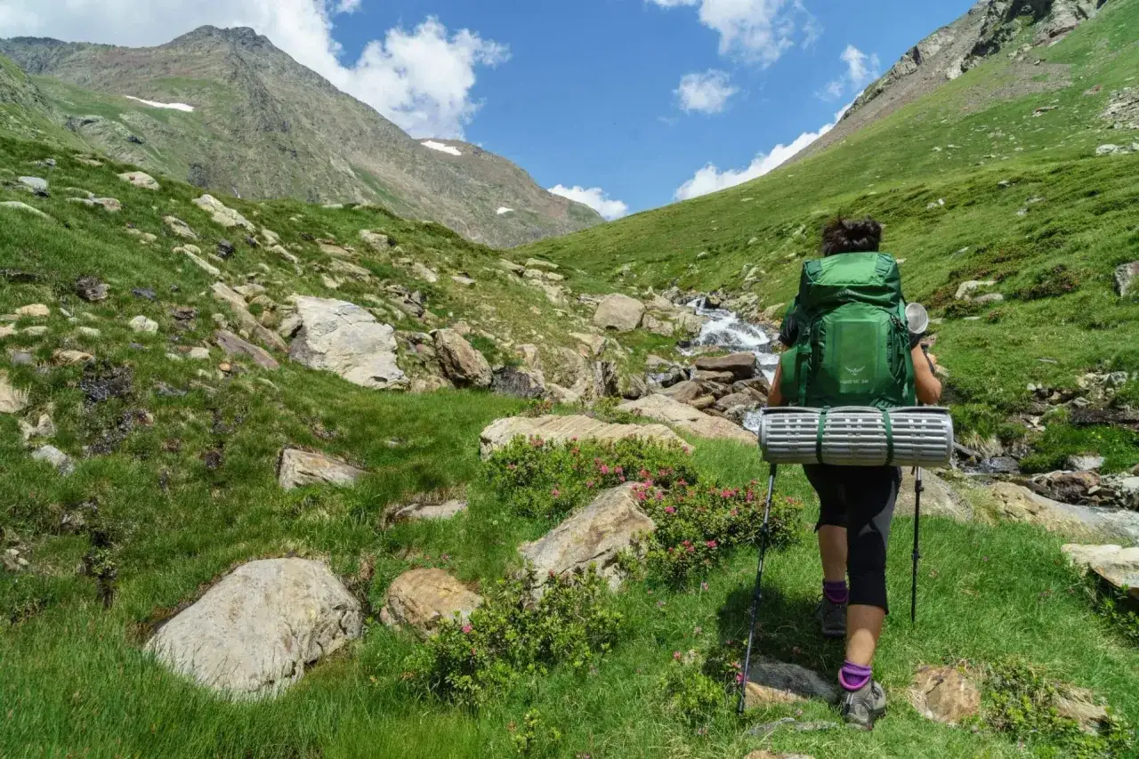 Une randonneuse avec un grand sac à dos progresse sur un sentier de montagne verdoyant, un tapis de sol enroulé. Un ruisseau serpente à ses côtés, le tout dans un paysage de trek pyrénéen spectaculaire.