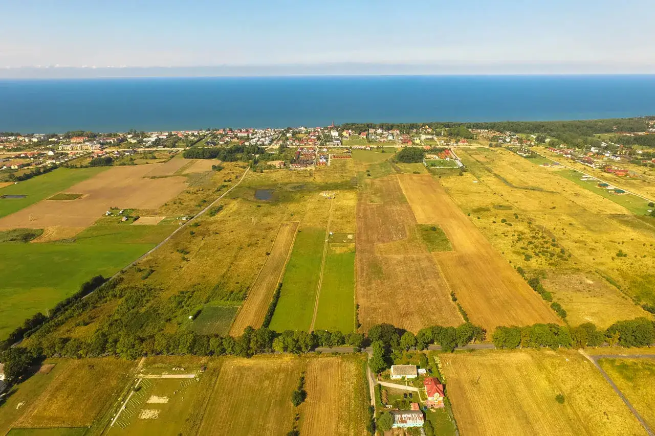 Widok z lotu ptaka na pola uprawne i zabudowania nad morzem. Rozważasz sprzedaż działki z warunkami zabudowy, a zastanawiasz się nad VAT?