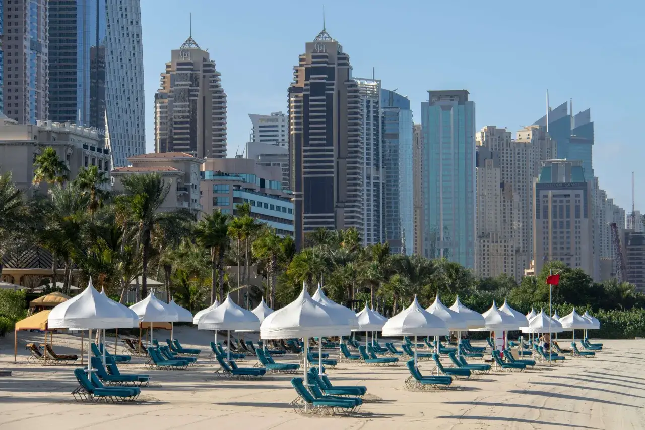 Piękna dubaj pogoda luty: pusta plaża z leżakami i białymi parasolami, w tle palmy i nowoczesne wieżowce.