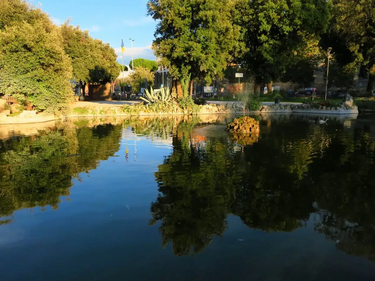 Un laghetto con riflessi verdi e blu, un'isola rocciosa con vegetazione e alberi che incorniciano un prato giardino a Viterbo.