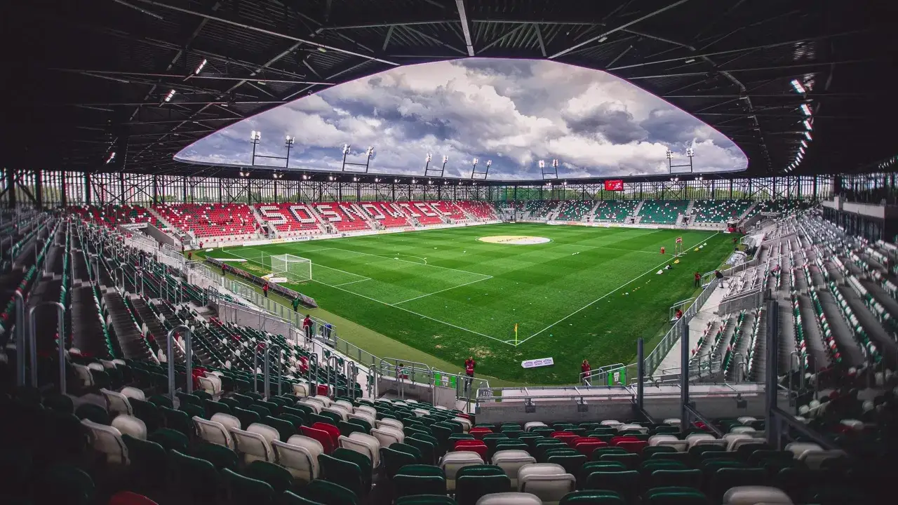 Stadion z zieloną murawą i trybunami w barwach zielono-czerwonych. Napis 