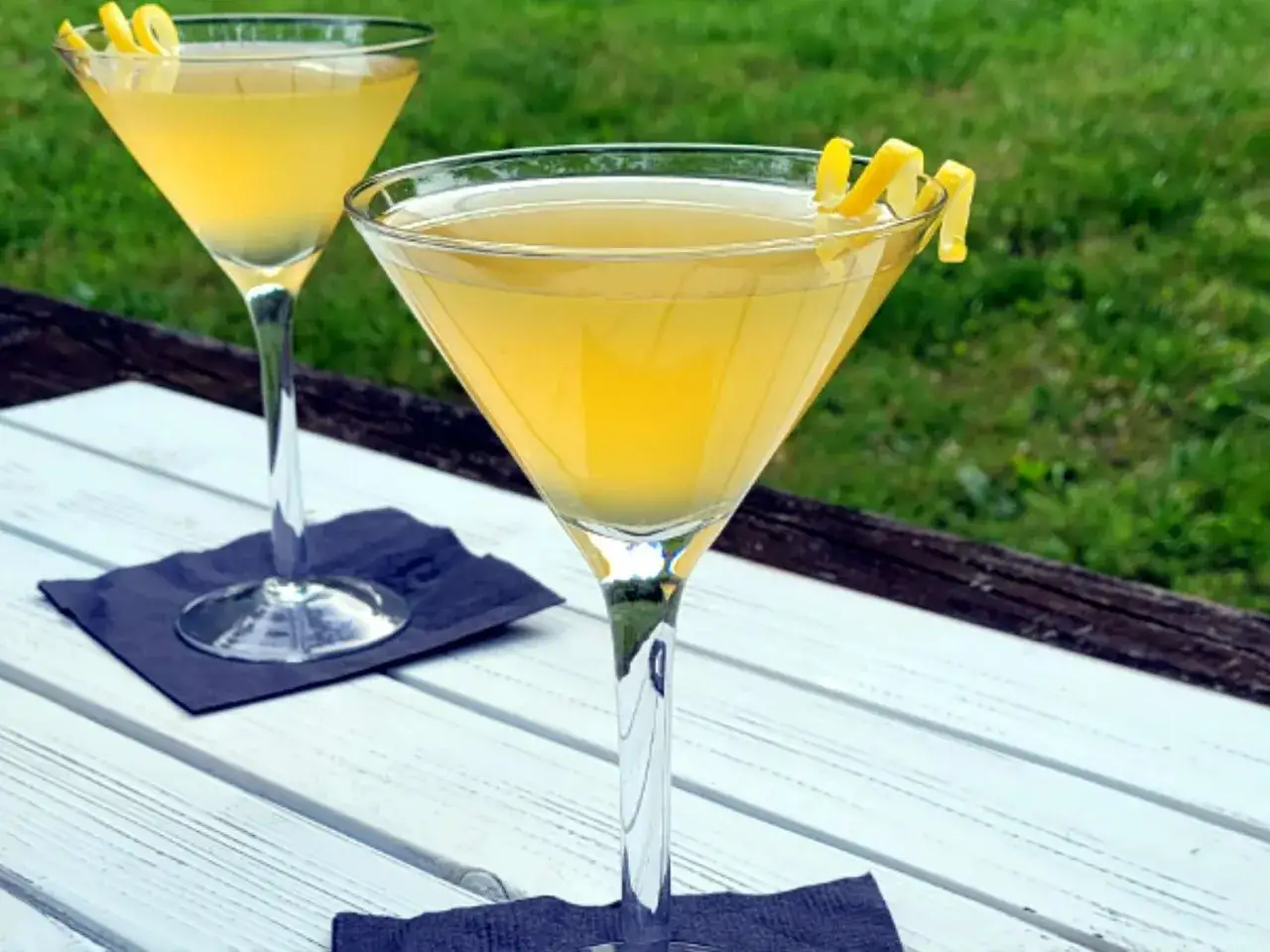 Dwa kieliszki z orzeźwiającym drinkiem passion fruit martini, ozdobione skórką cytryny, stoją na białym stole na tle zielonej trawy.