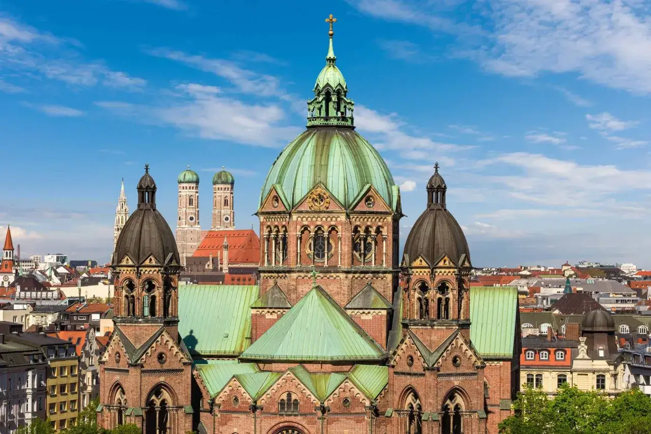 Prachtvolle Kirche mit grüner Kuppel und Türmen in **Lehel, München**. Im Hintergrund ragen die Türme des Münchner Doms empor.