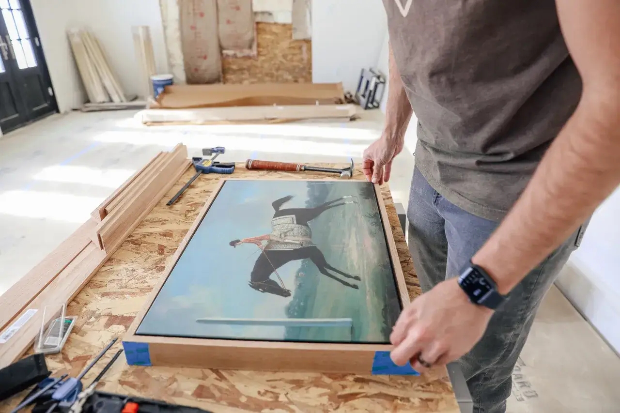 Hombre ajustando un lienzo con una pintura de un jinete a caballo en un marco de madera. Herramientas y molduras de madera listas para como enmarcar un lienzo.