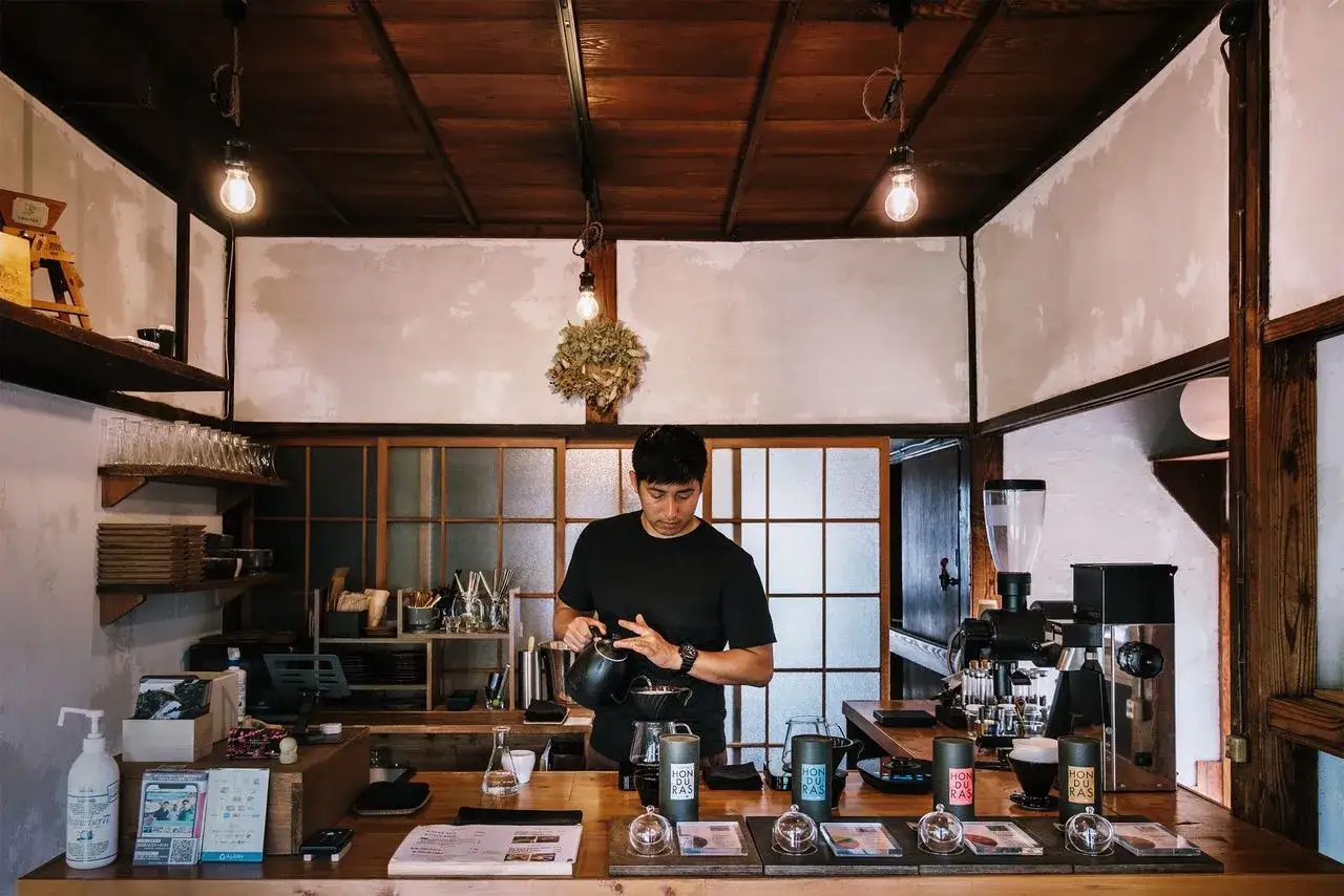 Barista prepara un caff&egrave; giapponese in un locale accogliente con soffitto in legno e luci soffuse.